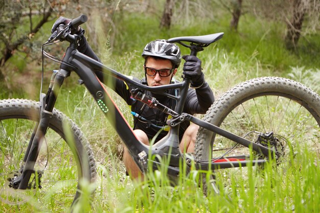 Disparo sincero al aire libre del joven jinete concentrado en equipo de protección sentado en el césped frente a su bicicleta eléctrica rota, tratando de descubrir cuál es el problema. Hombre comprobando e-bike antes cycli