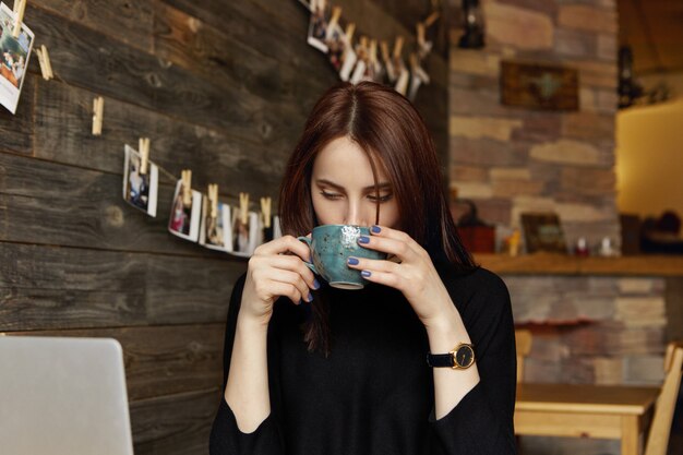 Disparo sincero de una bella mujer morena independiente en ropa negra tomando café o té de una taza grande durante un pequeño descanso mientras trabajaba remotamente en el café, sentado frente a la computadora portátil abierta