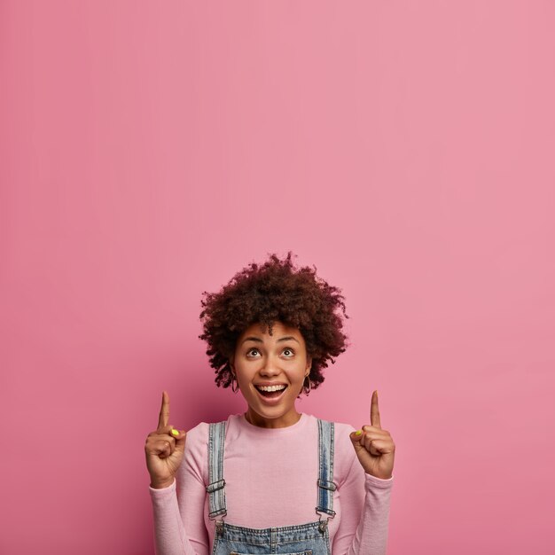 Disparo vertical de una alegre mujer étnica con puntos de pelo afro arriba, demuestra un impresionante espacio de copia, tiene una expresión de rostro alegre, muestra dientes blancos, se viste informalmente, promociona un artículo en un centro comercial o tienda