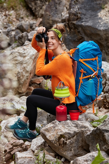 Disparo vertical de alegre viajero sentado en las rocas, hace maravillosas fotos a través de la cámara, hace café en la estufa de camping, viste un jersey naranja