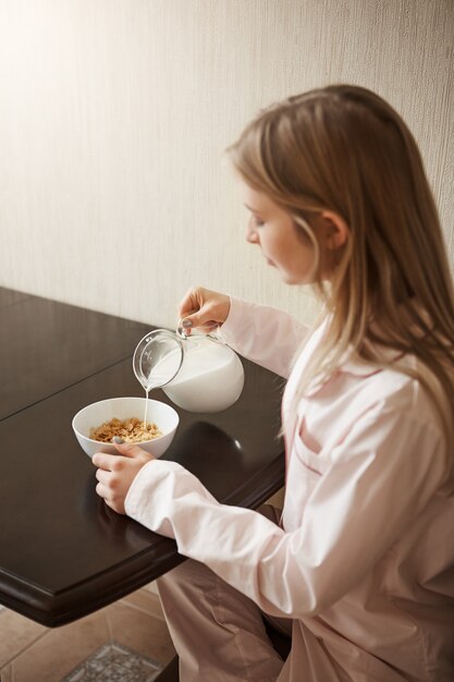 Disparo vertical de la atractiva hija rubia sentada en un acogedor pijama en la cocina, vertiendo leche en un tazón con cereales, desayunando sola, preparándose para estudiar en la universidad