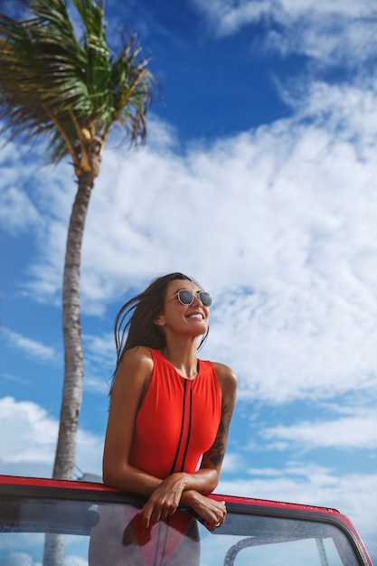 Foto gratuita disparo vertical de una atractiva modelo femenina en un traje de baño rojo, se para en el coche, se apoya en el parabrisas de un coche y sonríe.