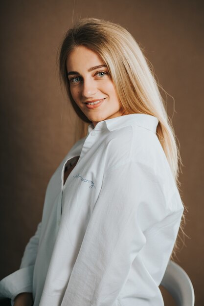 Disparo vertical de una atractiva mujer rubia con una camisa blanca posando sobre una pared marrón