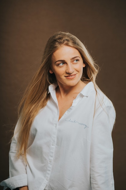 Disparo vertical de una atractiva mujer rubia caucásica con una camisa blanca posando sobre una pared marrón