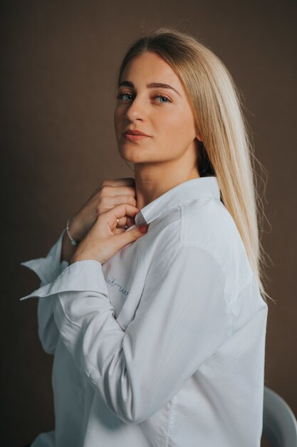 Disparo vertical de una atractiva mujer rubia caucásica con una camisa blanca posando sobre una pared marrón