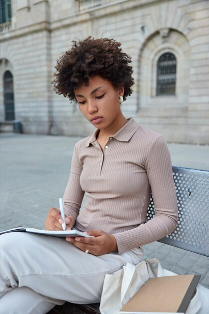 Disparo vertical de la autora seria toma notas en el bloc de notas para escribir posturas de artículos creativos en un banco en la antigua ciudad vestida con ropa casual