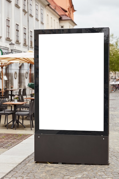 Disparo vertical de cartelera en blanco se encuentra en pavemenet contra el fondo de la ciudad cerca de la cafetería al aire libre