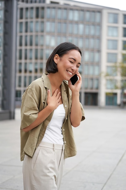 Foto gratuita disparo vertical de una chica asiática con estilo sonriente teniendo una conversación telefónica y caminando por la calle yo