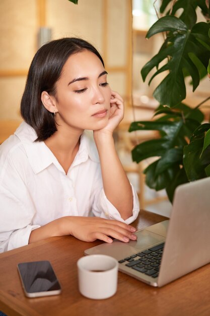 Disparo vertical de una chica asiática que parece cansada en la pantalla del portátil aburrida de trabajar sentada en un café con sueño