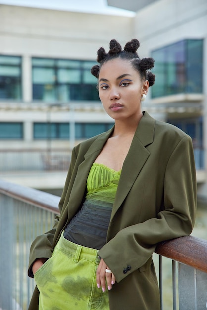 Foto gratuita disparo vertical de una chica milenaria seria con peinado de moño vestido con ropa verde de moda posa en el puente al aire libre contra el fondo borroso de la ciudad estilo de vida de la gente y concepto de vida urbana