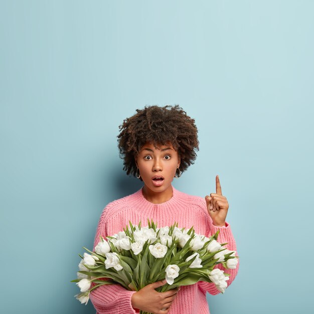 Disparo vertical de una dama afroamericana estupefacta apunta arriba con el dedo índice, ha contenido la respiración, sostiene un bonito ramo de tulipanes blancos de primavera, usa un jersey rosa casual, aislado contra la pared azul