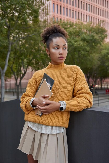 Disparo vertical de una estudiante pensativa y rizada sostiene una tableta digital y un cuaderno que va a la universidad usa un suéter amarillo de punto y una falda enfocada al aire libre contra el fondo urbano