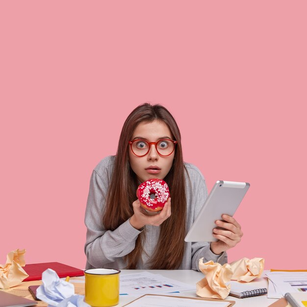 Disparo vertical de un estudiante universitario asustado que mira a través de las gafas, sostiene una deliciosa rosquilla en la mano, lleva un panel táctil actualizado, tiene muchos documentos en la mesa