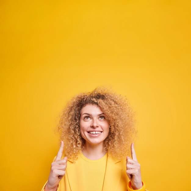 Disparo vertical de feliz mujer de pelo rizado apunta con el dedo hacia arriba muestra una oferta increíble con sonrisas desde arriba con dientes