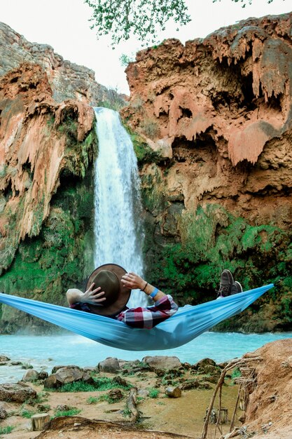 Disparo vertical de un hombre acostado en una hamaca junto a una cascada que fluye hacia abajo desde una colina