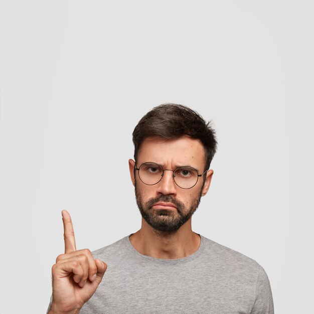 Disparo vertical de hombre joven con expresión de disgusto grave, barba densa, cabello oscuro, puntas con el dedo índice hacia arriba, vestido informal, aislado sobre una pared blanca. ¡Mira este!