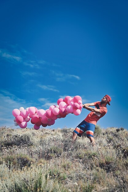 Disparo vertical de un hombre joven que lleva el manojo de globos de color rosa sobre una superficie de cielo azul