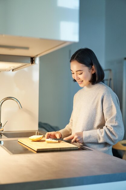 Disparo vertical de una joven asiática cocinando la cena haciéndose sandwitch sonriendo mientras está de pie