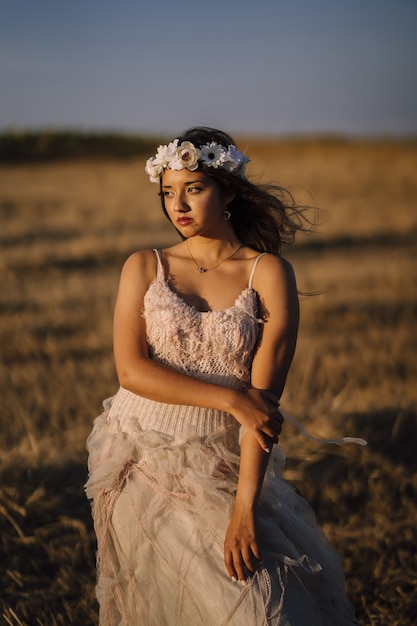 Disparo vertical de una joven mujer caucásica en vestido blanco y corona de flores blancas posando en un campo