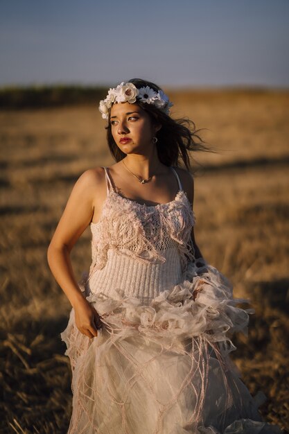 Disparo vertical de una joven mujer caucásica en vestido blanco y corona de flores blancas posando en un campo