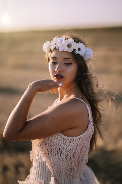 Disparo vertical de una joven mujer caucásica en vestido blanco y corona de flores blancas posando en un campo