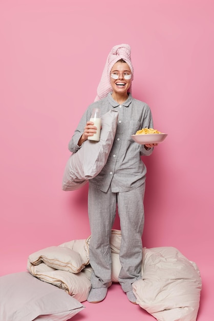 Disparo vertical de una joven positiva vestida con una toalla de pijama en la cabeza que tiene un desayuno saludable sostiene un tazón con copos de maíz y la leche se somete a procedimientos de belleza después de dormir disfruta de una gran mañana en casa