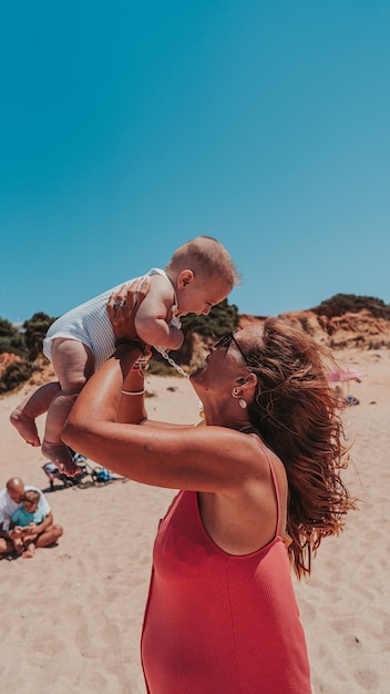 Foto gratuita disparo vertical de una mamá feliz abrazando a su bebé en una playa de arena