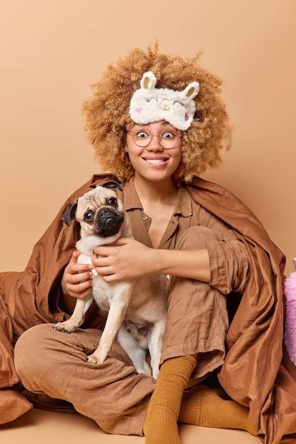 Disparo vertical de mujer alegre con cabello rizado y tupido vestido con pijama juega con perro pug vestido con pijama y antifaz para dormir aislado sobre fondo marrón Feliz dueño de mascota en el dormitorio yendo a dormir