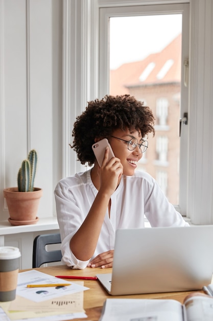 Disparo vertical de mujer alegre contable habla por teléfono móvil, tiene expresión positiva, trabaja con computadora portátil