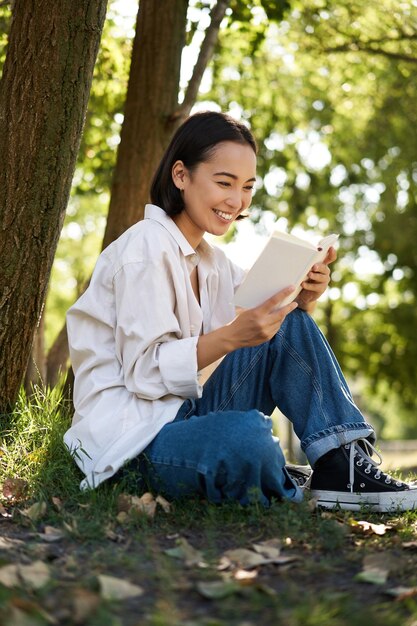 Disparo vertical de mujer asiática feliz relajándose al aire libre en el parque leyendo su libro y sentada bajo un árbol