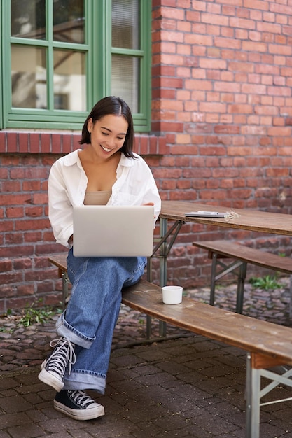 Disparo vertical de una mujer asiática que trabaja en un café al aire libre sentado con una computadora portátil en un banco y bebiendo café