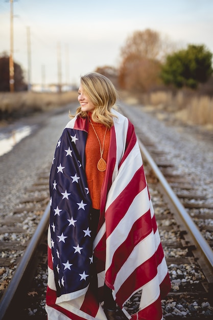 Disparo vertical de una mujer con la bandera americana sobre sus hombros de pie sobre el ferrocarril