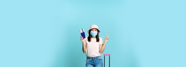 Foto gratuita disparo vertical de mujer coreana feliz con máscara médica con boletos de avión y pasaporte de pie con ingenio