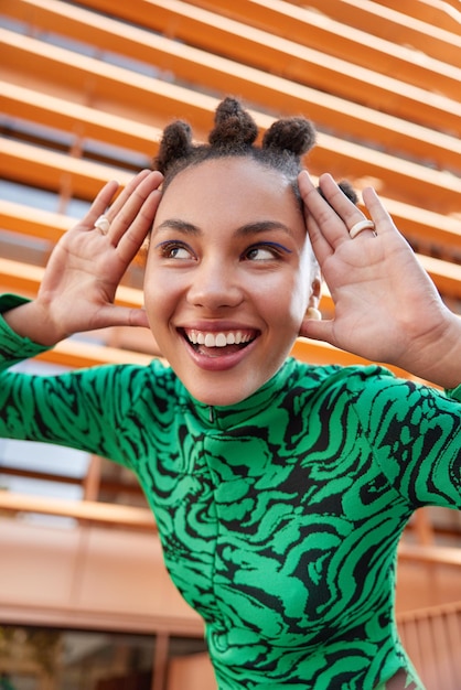 Foto gratuita disparo vertical de mujer joven feliz con peinado de moño mantiene pams cerca de la cara sonríe con dientes aplica delineador de ojos azul usa poses de puente verde al aire libre contra un fondo borroso tiene un estado de ánimo juguetón