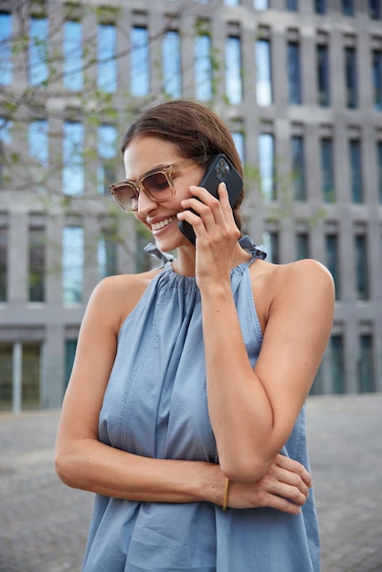 Disparo vertical de mujer joven feliz tiene conversación telefónica discute algo con amigo tiene humor optimista viste ropa elegante