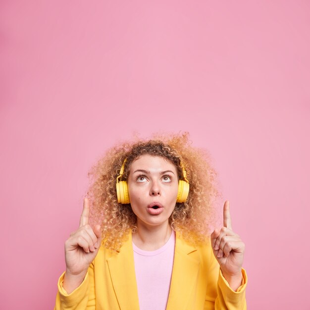 Disparo vertical de una mujer de pelo rizado sin palabras con expresión de asombro que indica hacia arriba, demuestra una oferta inesperada, lleva auriculares inalámbricos en las orejas y disfruta de la lista de reproducción favorita