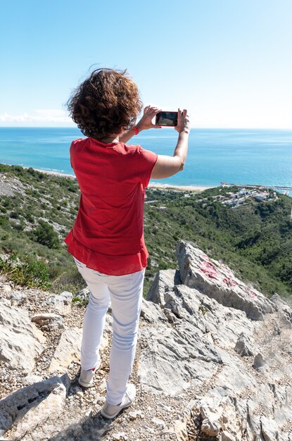 Disparo vertical de una mujer de pie sobre las rocas y fotografiando el hermoso mar