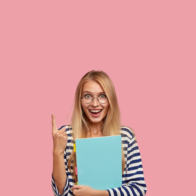 Disparo vertical de mujer rubia alegre con expresión positiva, señala con el dedo índice hacia arriba, lleva el libro de texto azul, muestra el espacio libre
