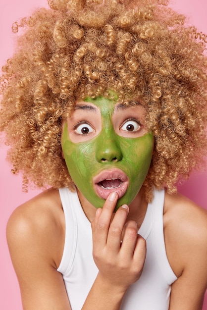 Disparo vertical de una mujer sorprendida y asustada con el pelo rizado mira fijamente los ojos saltones mantiene la boca abierta por el impacto aplica una máscara nutritiva verde en la cara para el tratamiento de la piel posa en el interior con una camiseta informal