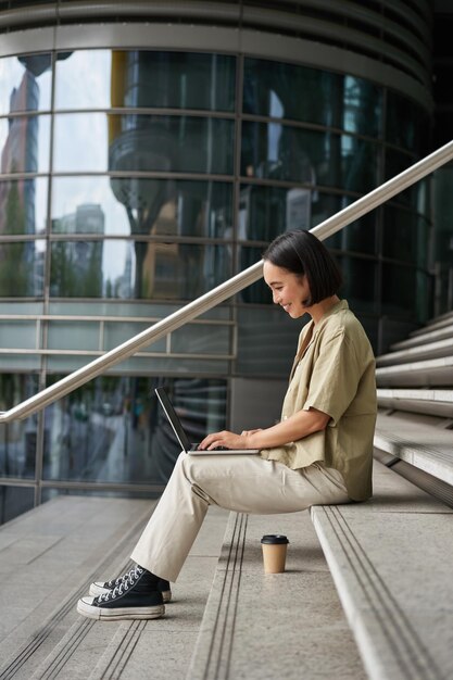 Disparo vertical de niña asiática sonriente con café usa laptop al aire libre mujer joven con computadora outsi