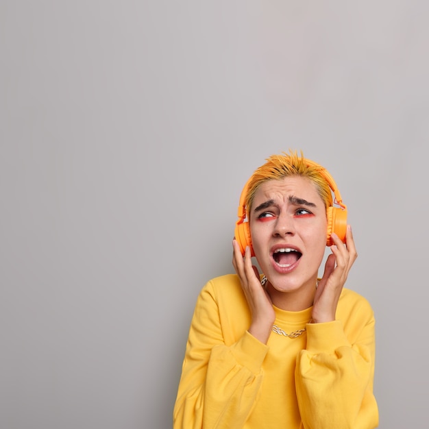 Disparo vertical de optimista chica de pelo amarillo con apariencia inusual maquillaje brillante escucha música en auriculares inalámbricos canta canciones a lo largo aislado sobre pared gris
