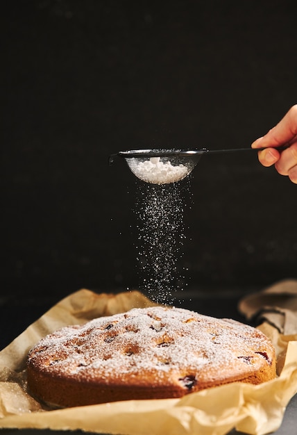 Disparo vertical de un pastel de cerezas con azúcar en polvo e ingredientes en el lateral sobre un fondo negro