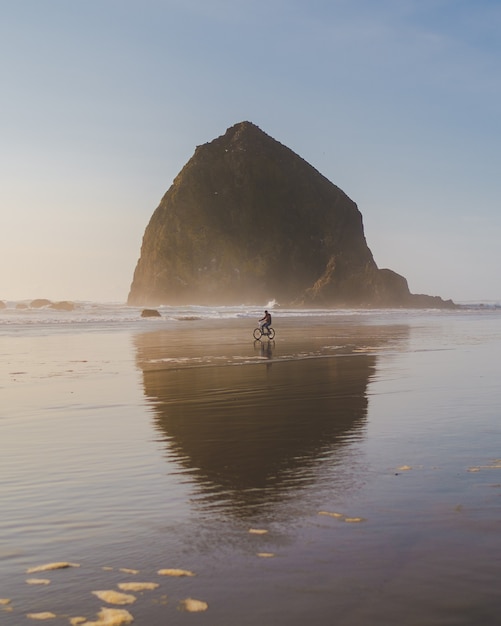 Foto gratuita disparo vertical de una persona en bicicleta a la orilla del mar con una gran roca en la distancia