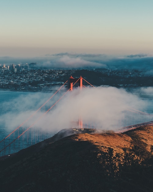 Foto gratuita disparo vertical del puente golden gate detrás de las nubes con edificios en la distancia