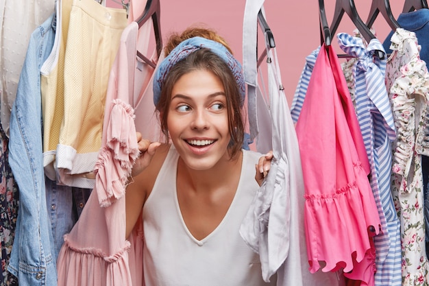 Foto gratuita disparos en la cabeza de la encantadora y juguetona joven europea jugando al escondite con su hermano pequeño, escondiéndose entre un montón de ropa colgada en el estante de su armario. concepto de compras y consumismo