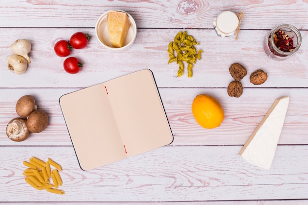 Disposición del ingrediente de pasta y diario abierto en blanco en el escritorio de madera