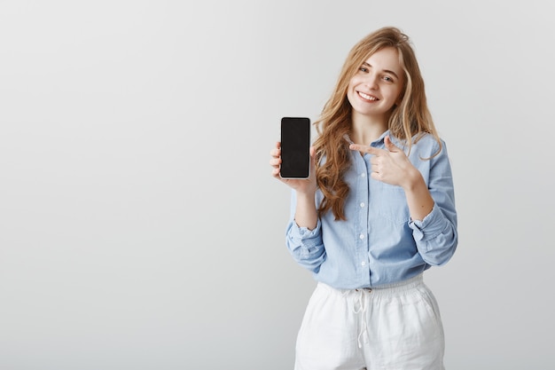 Dispositivo super útil. complacida y guapa estudiante con cabello rubio y camisa de cuello azul, mostrando un smarpthone negro y apuntando al dispositivo con el dedo índice, ofreciendo comprar un artículo