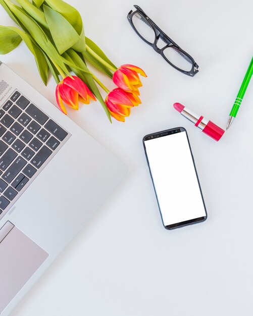 Dispositivos electrónicos cerca de flores, lápiz labial y anteojos.