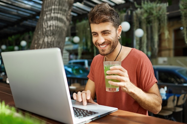 Distancia social alegre chico barbudo con amigos, videollamadas con computadora portátil mientras está sentado en el área de café al aire libre
