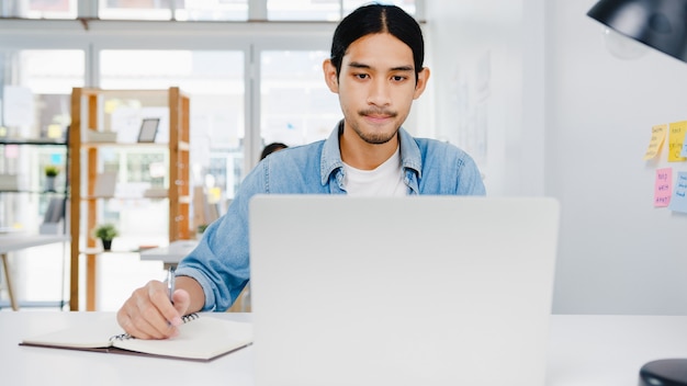 Distanciamiento social del empresario de Asia en una nueva situación normal para la prevención de virus mediante la presentación de una computadora portátil a un colega sobre el plan en la videollamada mientras trabaja en la oficina en casa. Estilo de vida después del virus corona.